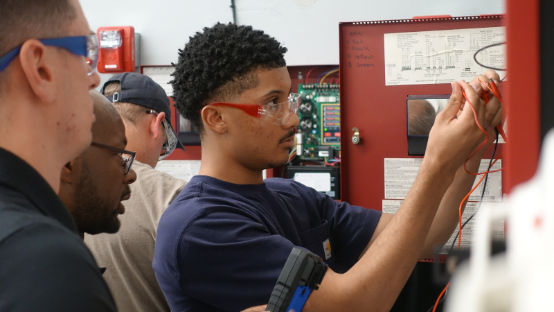 A young man wearing safety glasses works in a Fire Alarm panel while others watch.