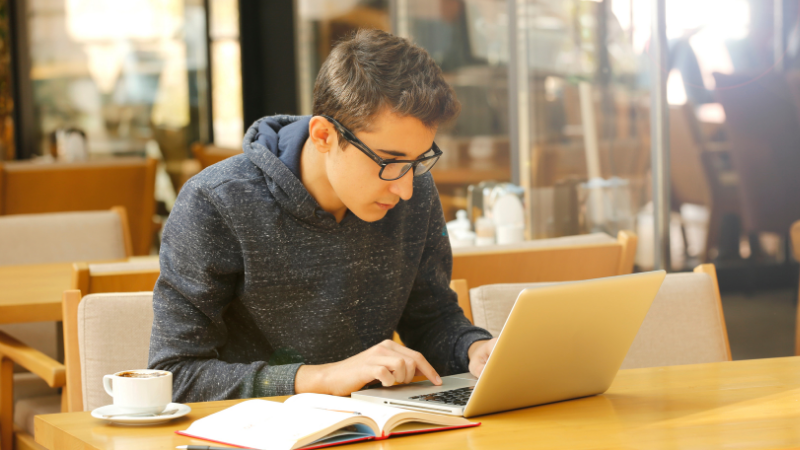 A man with a blue sweatshirt and glasses studies on his laptop in a coffee shop.